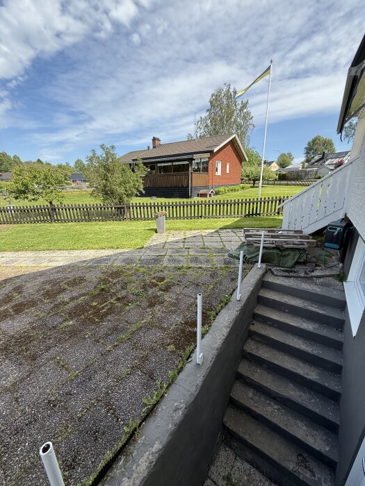 Vy över en trädgård med gräsmatta och staket, trappa ned till plattbeläggning, rött hus och flaggstång i bakgrunden.