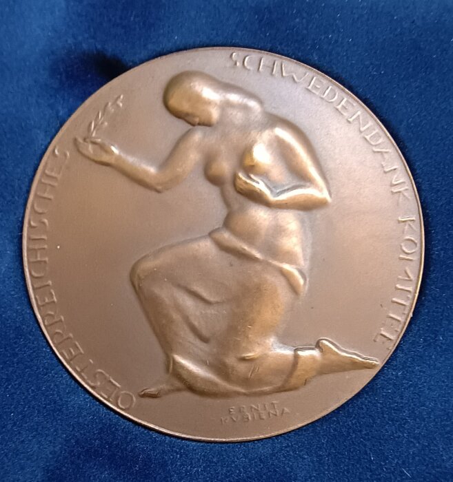 Bronsmedalj med relief av knäböjande figur som håller en olivkvist, omgiven av texten "ÖSTERREICHISCHE SCHWEDENDANK-KOMITEE".