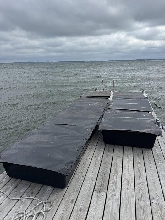 Flytbrygga under konstruktion med svart plasttäckta pontoner vid en brygga, med molnig himmel och sjö i bakgrunden.