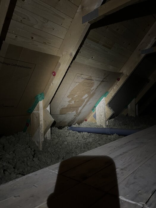 Bilden visar en vindssektion med träbalkar och isolering. Det finns en skuggad fläck på takpanelen som kan indikera tidigare vattenläckage.