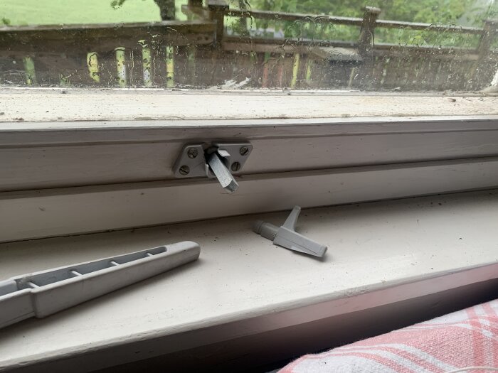 Trasigt fönsterhandtag liggande på fönsterbrädan med utsikt genom smutsigt fönster.