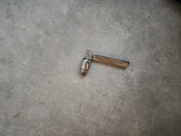 Trasigt dörrhandtag i metall ligger på grått golv, handtaget har gått av vid den del som ska fästas i altandörren.