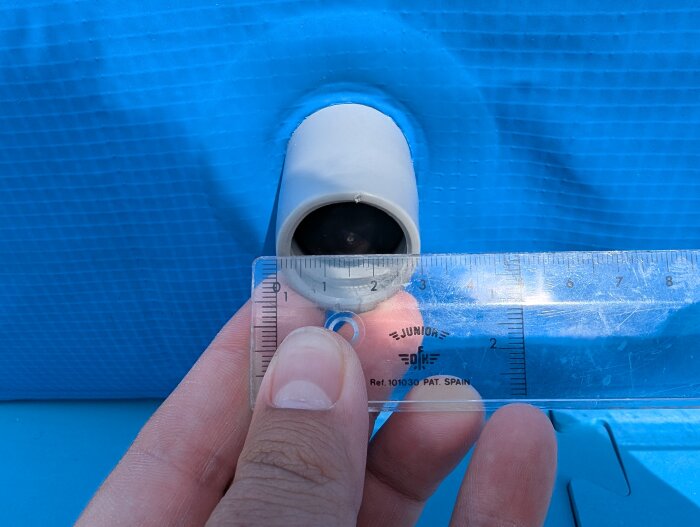 Blå barnpool med gummiuttag och en hand som håller en linjal mot öppningen. Uttaget mäts och har en diameter på 30 mm.