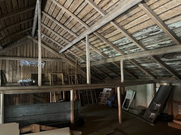 Ett snedtak med träkonstruktion och loft, flera stödjande bjälkar och brädor. Olika föremål lagrade på loftet i ett oinrett utrymme.