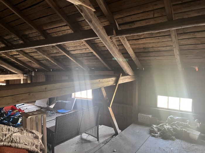 Loft med träbjälkar och takstolar, fönster ljusar upp rummet. Möbler och föremål syns längs väggarna.