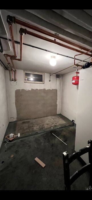 Pannrum med betonggolv och väggar, synliga oljefläckar och kopparrör i taket, förbereds för omvandling till tvättstuga.