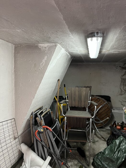 Källarrum med stolar staplade mot en vit vägg, lysrör i taket och flera föremål på golvet inklusive en trädgårdsslang och en grön säck.
