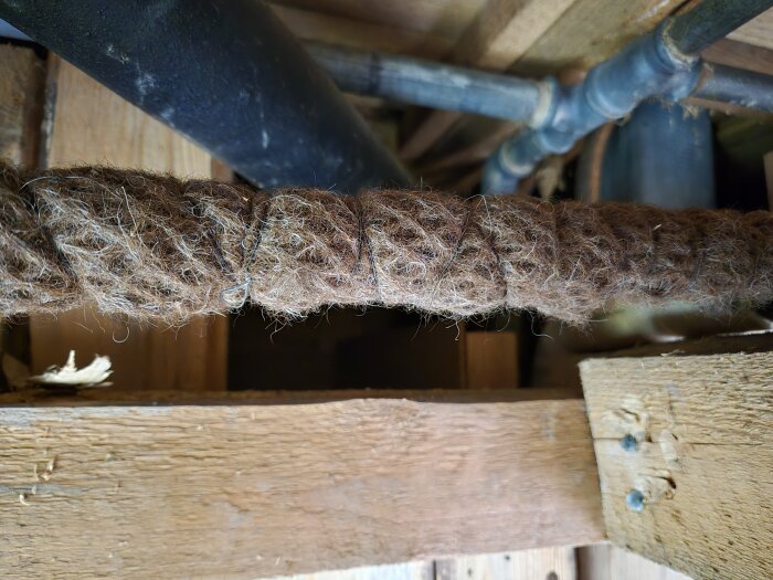 Brunt isoleringsmaterial i ett snurrat mönster, möjligtvis asbest eller ett annat material, synligt ovanpå träbjälkar och rör i en byggnad.