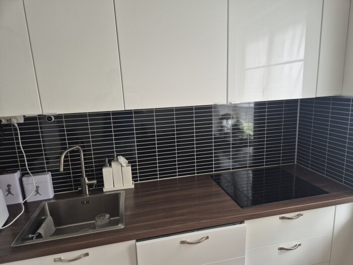 Modernt kök med svarta kakelplattor som stänkskydd, vita skåp och träbänkskiva med diskho och induktionshäll.