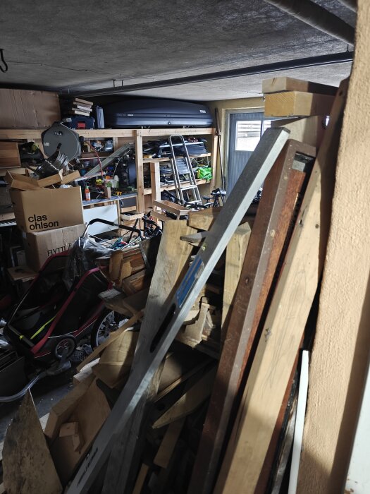 Rörigt garage med verktyg, träbitar och en hylla fylld med diverse föremål, samt en takbox och en barnvagn.