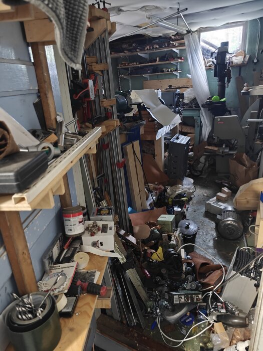 Ett rörigt garage och verkstad med verktyg, maskiner och material spridda över hyllor och golv, visar pågående byggprojekt och organisering.