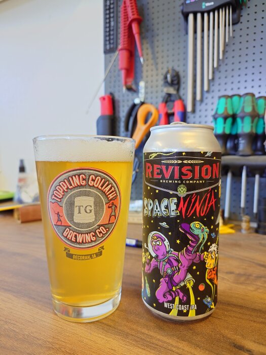 Ett glas öl från Toppling Goliath Brewing Co. bredvid en burk Revision Brewing Company Space Ninja IPA på en arbetsbänk med verktyg i bakgrunden.