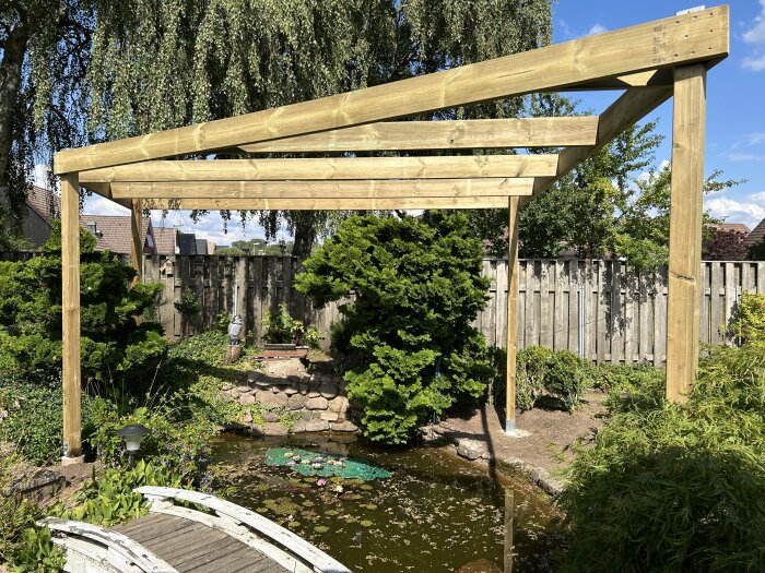 Pergola byggd över en damm med omgivande stenar och grönska, i en trädgård med staket och träd i bakgrunden.