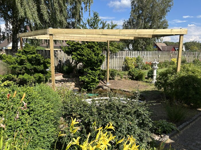 Pergola byggd över en damm i en trädgård, omgiven av buskar och staket i bakgrunden under en solig dag.