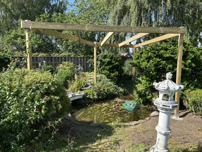 Pergola byggd över en trädgårdsdamm omgiven av grönska, med en stenlykta i förgrunden.