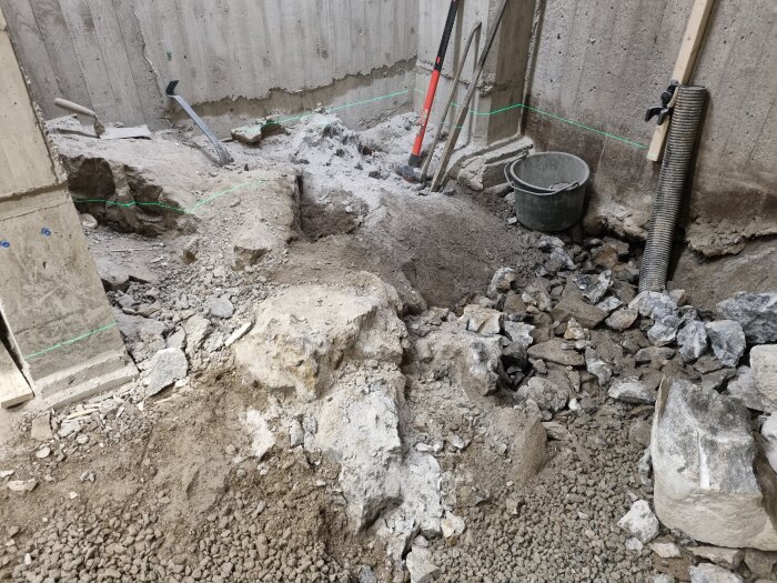 Byggarbetsplats med grävt golv och stenar, spade, hacka, och hink synliga.
