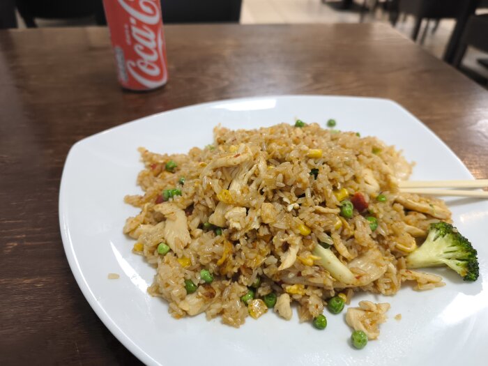 Stekt ris med kyckling, ärtor, broccoli och mais på en vit tallrik. I bakgrunden syns en burk Coca-Cola på ett brunt bord.