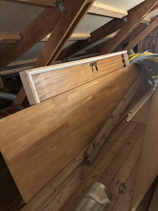 En komplett dörr med karm, lutad mot takbjälkar på en vind, omgiven av diverse byggmaterial och ett gult rör.