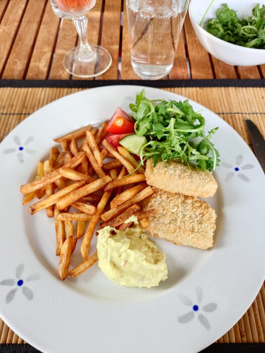 Tallrik med fisk, pommes frites, remouladsås, tomat och sallad, serverad på ett bord med dryck i glas och en skål med mer sallad.