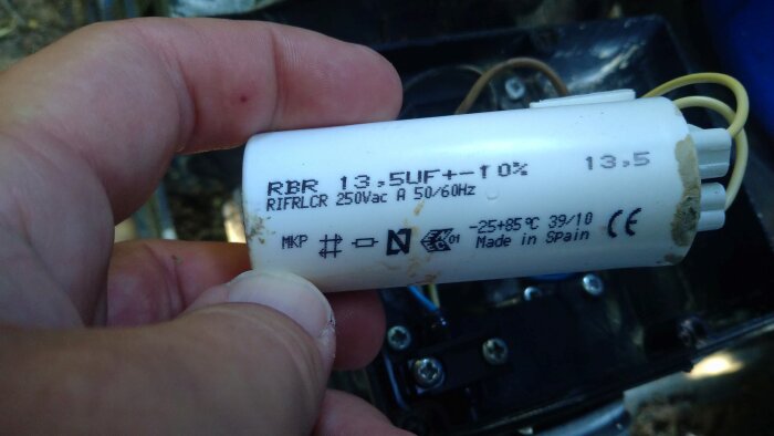 En hand håller en vit startkondensator märkt 13.5 uF och 250 Vac, del av diskussion om ersättning av poolpumpkomponent.