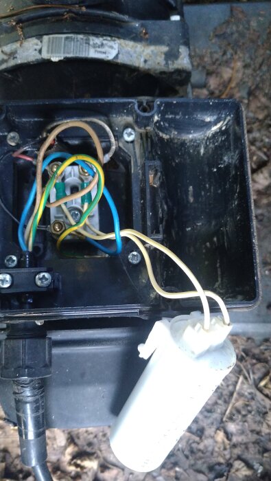 En öppen elektrisk kopplingsbox med färgade ledningar och en vit kondensator, omgiven av smuts och löv.