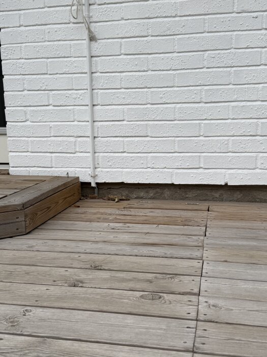 Vita tegelväggar med synliga sprickor i sockeln på ett 70-talshus, omgivna av trätrall på marknivå.