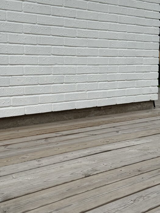 Vitt tegelvägg ovanför ett trädäck med sprickor i betonggrunden.