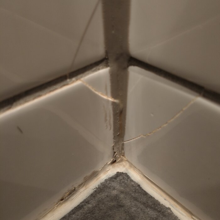 Spruckna kakelplattor i ett hörn av ett badrum med mörk fog och ljusgrått golv.