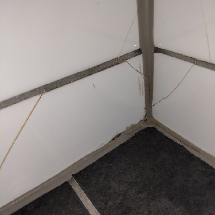 Sprickor i vitt kakel i badrumshörn med mörk fog och golvplattor.