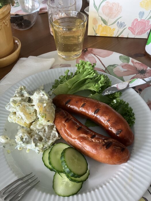 Grillad chorizo, hemmagjord potatissallad och gurkskivor på en tallrik. Bakgrund med glas dryck och blommigt servettställ.