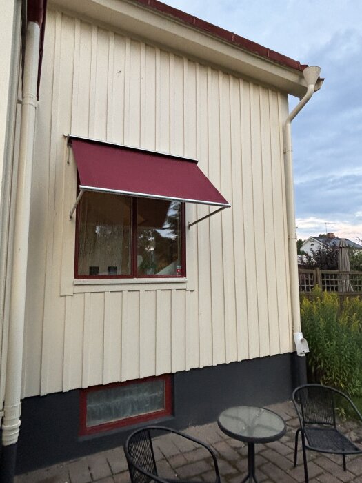 Gult trähus med oxydröda fönster och röd markis, gråsvart sockel. Utemöbler i svart på stenlagd yta.