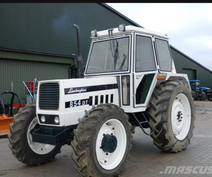 En vit Lamborghini 854 DT traktor står vid en byggnad, med stora hjul och en fyrkantig hytt.