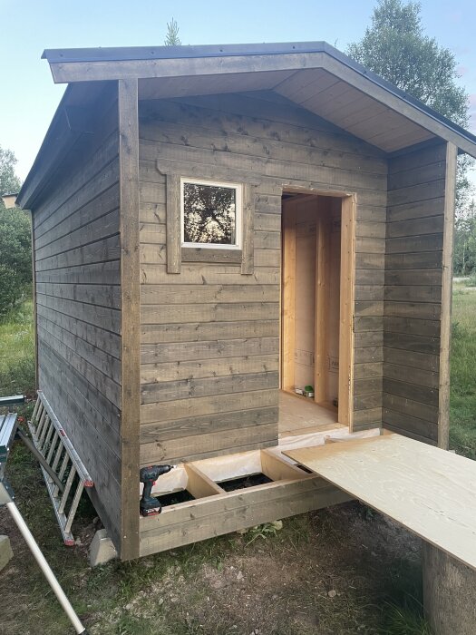Litet trähus under konstruktion med oisolerat golv och öppning för dörr, fönster på väggen. Stege och verktyg syns vid sidan.