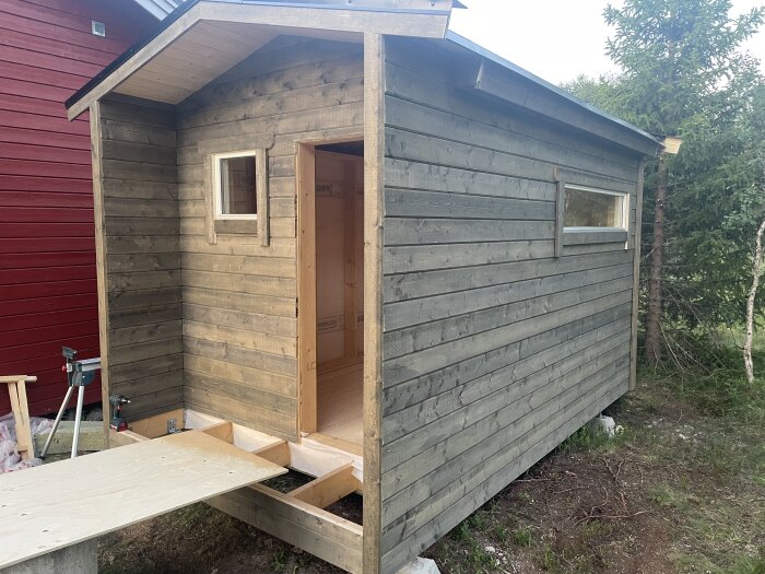 Litet grått trähus med öppnad dörr och två fönster, beläget bredvid ett rött hus och träd i bakgrunden, träpanel på marken.