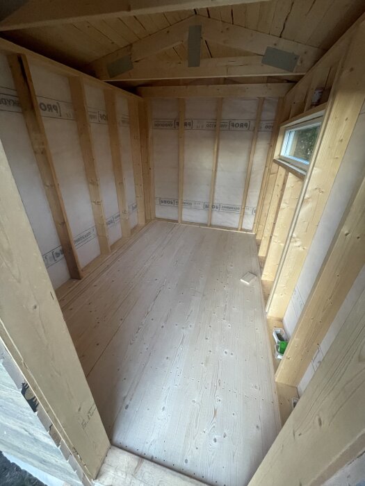 Inre del av ett träbyggnadsprojekt med isolerade väggar och nylagt golv, fönster synligt till höger.