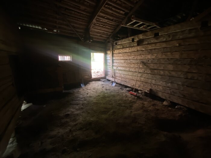 Mörk lada med jordgolv och träväggar, ska omvandlas till garage. Verktyg syns på golvet och ljus strömmar in genom en dörröppning.