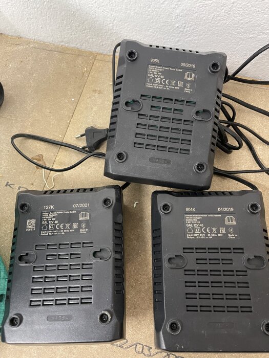 Tre svarta 12V-laddare från Robert Bosch Power Tools med etiketter och sladdar, liggande på en träyta.