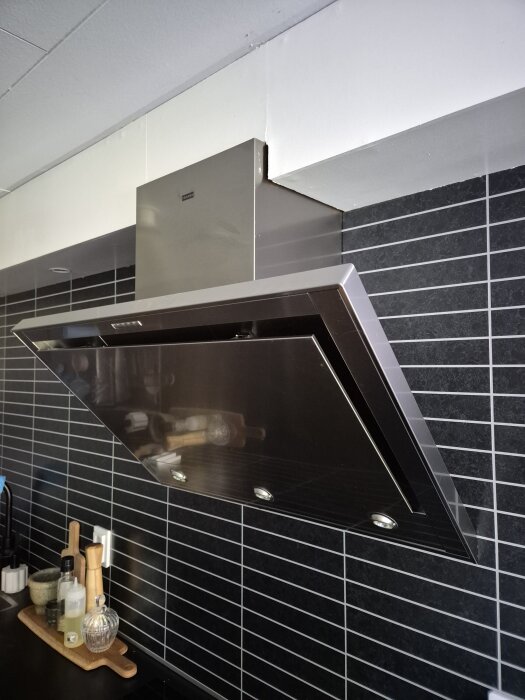 Köksfläkt monterad ovanför spis med svart kakel i bakgrunden.