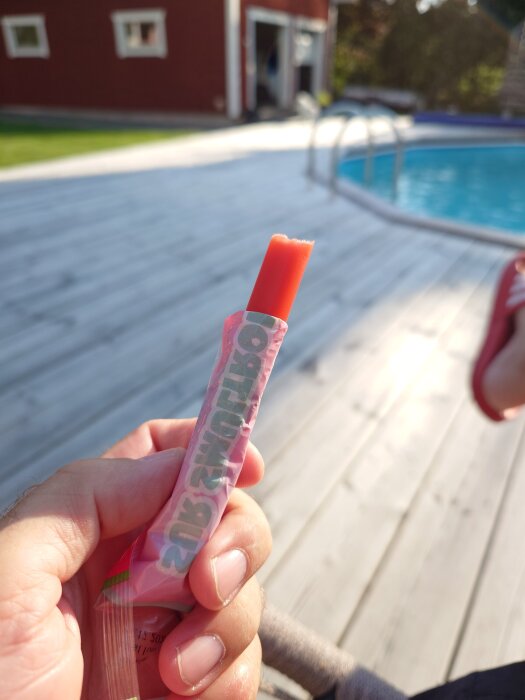 En hand håller en halväten isglass vid en pool på en träaltan, med en röd byggnad i bakgrunden.