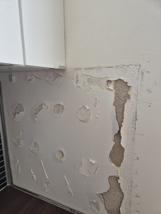 Vägg med skadad puts bredvid köksskåp, flera områden med avskalad färg och exponerad yta, tecken på renoveringsbehov.