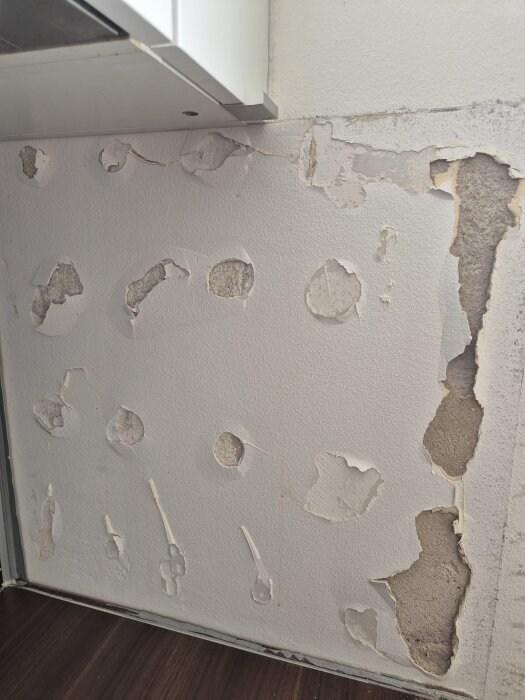 Vägg med avskavd färg och skador, exponerar betong och gips. Tecken på vatten- eller fuktskada ovanför en bänkskiva i kök.