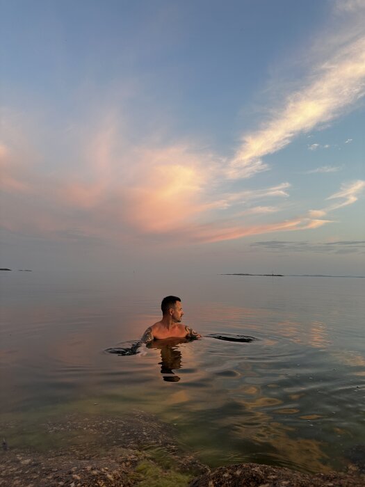 Person badar i en sjö vid solnedgången, med färgglada moln på himlen och spegelblanka vattnet.