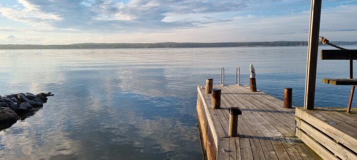 Träbrygga vid en sjö med klara vattenytor och en trappa för bad, skuggor tydliga i morgon- eller eftermiddagssol.
