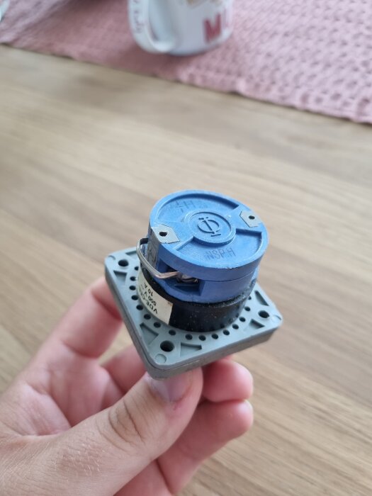 En hand håller en återfjädrande tryckknapp för start/stopp, liknande de som används på fartyg. Bakgrunden visar en bordsyta och en mugg.