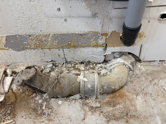 Stort rör synligt under golv och vägg i kök, avslöjat efter borttagning av skåp och golv. Rörledning omgiven av betong och damm.