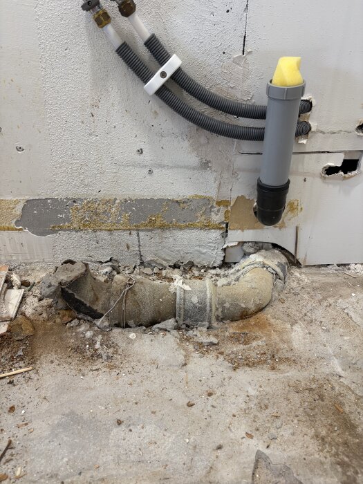 Ett stort rör sticker upp från golvet i ett kök, omgivet av slitna ytor och trasiga plattor, med ett hål i väggen ovanför.