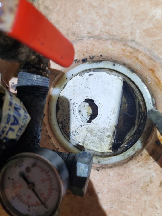 Vattenlås i tvättrum med synlig smuts och trasig tätning, omgiven av rördelar och mätare.