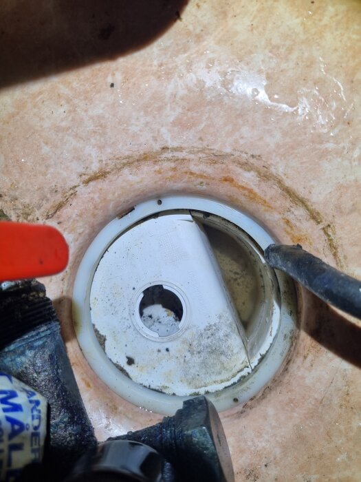 Närbild på ett smutsigt och delvis öppet vattenlås i ett tvättrum, omgivet av verktyg och tecken på korrosion i rören.