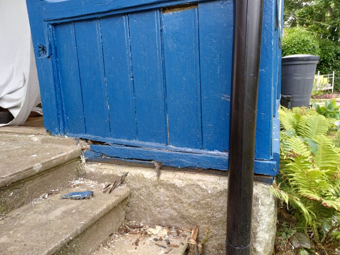 Blå glasveranda med rådnet træværk i bunden ved siden af en nedløbsrør og trapper.