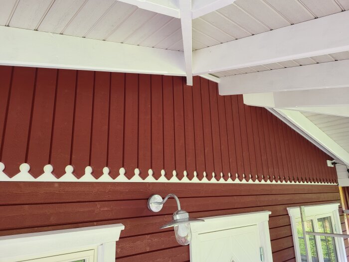 Röd träfasad med vit dekor vid takfoten, ovanför vita dörr- och fönsteromfattningar med synliga takstolar och utelampa.